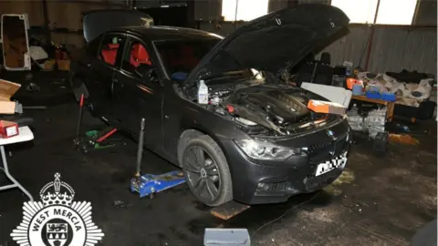 West Mercia Police A black BMW car with the hood open while it is elevated off of the ground inside a garage