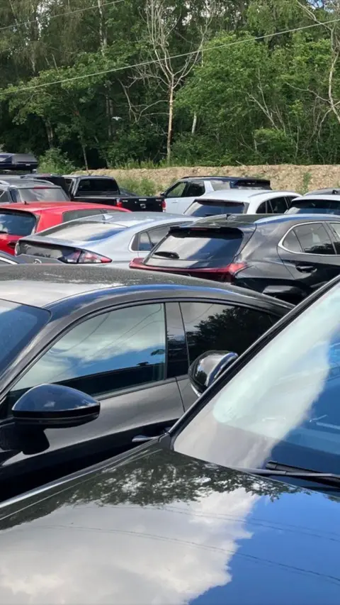 Lots of cars parked closely together with a fence and trees in the background