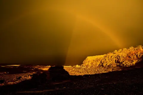John Cuthbert A rugged landscape glows under an intense golden-yellow sky, casting dramatic shadows across the ground. A faint rainbow arcs through the surreal light, blending softly into the horizon.