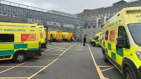Eight ambulances are parked outside Derriford Hospital and there are people walking past them.