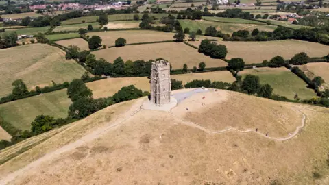 Getty Images A picture taken above Glastonbury Tor. Most of the grass in the foreground is dry and brown, while there are some greener fields and bushes in the background.