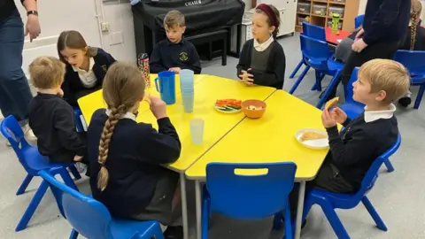 A mixed gender group of schoolchildren sit on blue plastic chairs around a yellow table, having breakfast. They wear dark blue jumpers with white polo shirts.