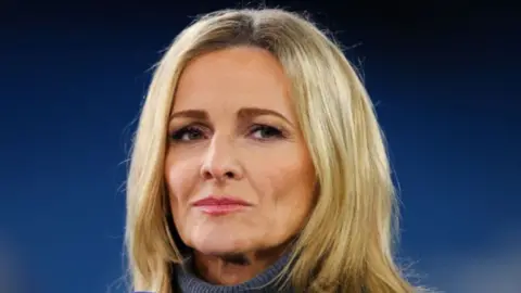 A blonde-haired woman with a serious face, wearing a grey turtleneck looking off into the distance.