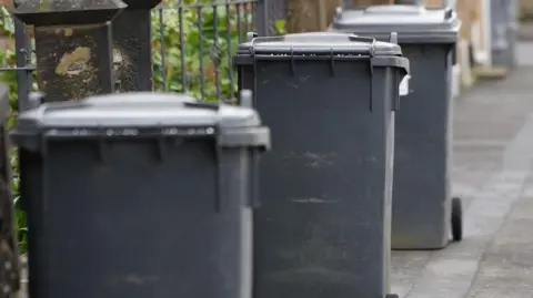 Black wheelie bins left out for on a street. There are three bins on the pavement. There is metal fencing behind the bins.