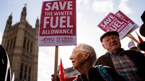 EPA Two elderly protesters hold red placards "save winter fuel allowance" Outside the Parliament House, which can be seen in the background. One is looking towards the left and the other is looking towards the camera wearing a flat cap and checked shirt.