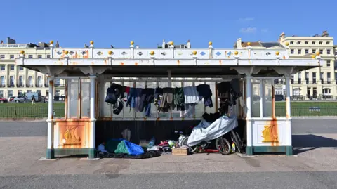 A rusty shelter on Hove seafront which has a homeless person's possessions inside. Including hanging clothes, bikes and a sleeping bag.