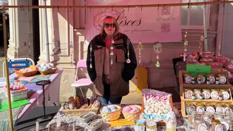 LDRS Cherie Bennett is wearing a large outdoor coat and shades. She's standing inside her market stall, which sells a range of confectionary and cakes.