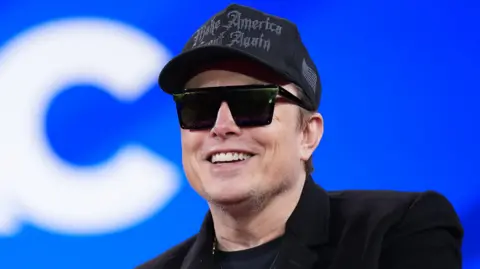 Elon Musk wears a black 'Make America Great Again' cap and sunglasses while smiling, on a blue backdrop.