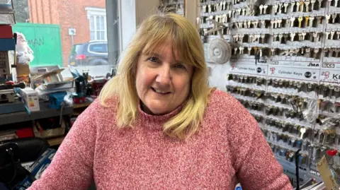 Martin Heath/BBC Karen Parisi with long blonde hair smiling at the camera and wearing a red and white sweater. There are racks of keys behind her and a window out onto the street, in which a green lorry can be seen.