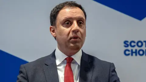 Anas Sarwar, who has short dark hair, at a press conference in front of a saltire sign. 
