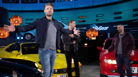 Flintoff with arts outstretched on the top Top Gear set, in front of Paddy McGuinness and Chris Harris and two cars