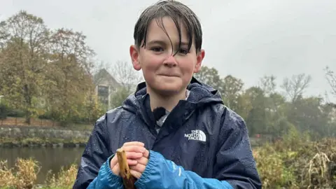 Joel is a young boy and is wearing a North Face blue rain jacket. He is resting his hands on a wooden staff and is wet from the rain. He is standing beside the River Kent 