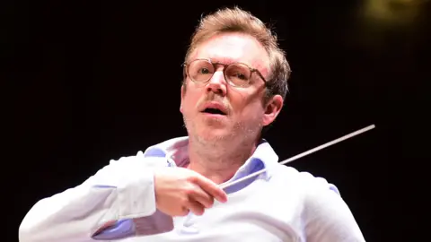 Daniel Harding in an open-necked white shirt with an arm raised holding a conductor's baton conducting the Orchestra Santa Cecilia of Roma in Bologna on 8 May 2026