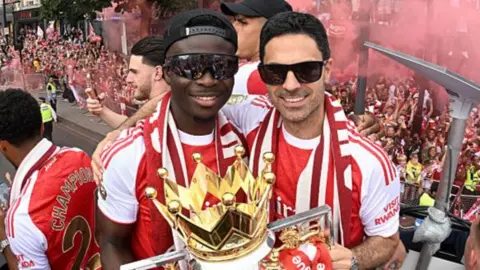 Bukayo Saka and Mikel Arteta, both in sunglasses and red and white team kit, with the Premier League trophy on Arsenal's bus parade