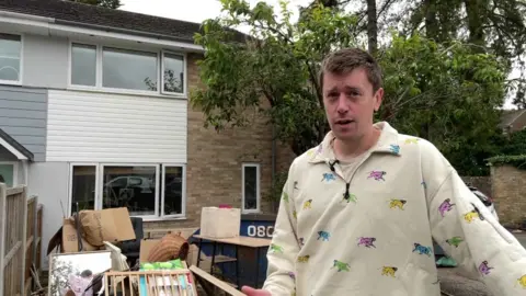 Will Capper wearing a beige jumper with monkeys on it in different colours his flooded family home in the background