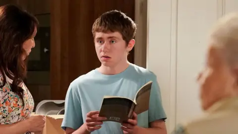 BBC/Jack Barnes/Kieron McCarron Barney Mitchell, played by Lewis Bridgeman, is stood holding a book and dressed in a blue t-shirt. He is stood next to Honey Mitchell and Big Mo.