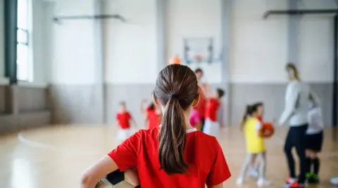 Getty Images A young girl with a football under her arm facing away from the camera with a PE lesson going on in the background