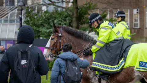 West Midlands Police Two horses ridden by people in yellow reflective jackets and black riding helmets next to two people in dark coats