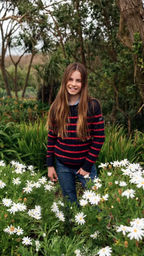 Kensington Palace Photograph shows Charlotte wearing a black and red striped jumper and blue denim jeans, standing in a flowery field while smiling at the camera.
