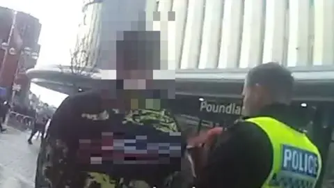 A police officer on the right of the image is speaking to a man in a black jumper with light green patterns and a logo on it. The logo is blurred, and so is the man's face. In the background is a large building which appears to be a multistorey car park, with a poundland shop on the ground floor. They are on a city centre street.