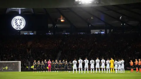 SNS Two rows of football players applauding in front of a dark stadium filled with fans.