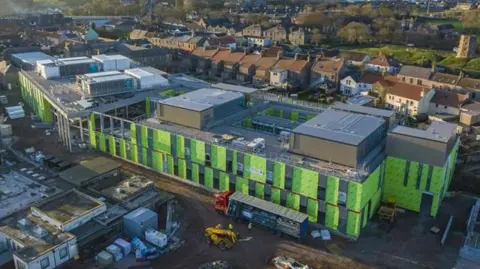 An aerial view of the new Berwick hospital mid construction. Green panelling has been paced over the sprawling hospital building while work is under way. An excavator and a large lorry are parked outside. There are rows of houses behind it.