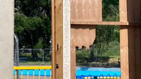 A wooden fence has some of its panels removed due to damage. Behind it you can see a black railing and a yellow railing higher up and a blue turquoise outdoor small pool behind it, with metal fencing around it. Trees in a park can be seen behind it.