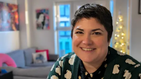 BBC Dee Montague-Coast is looking at the camera smiling. She has short black hair, and is wearing a green cardigan with a sheep pattern on. There is a Christmas tree in the background, in her Newport flat.