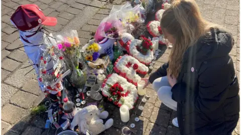 Ms Beeton kneels in front of the memorial at the site where Mr Dunn lost his life. Flowers, teddys, photos and bottles of his favourite cherry coke are some of the items left at the site.