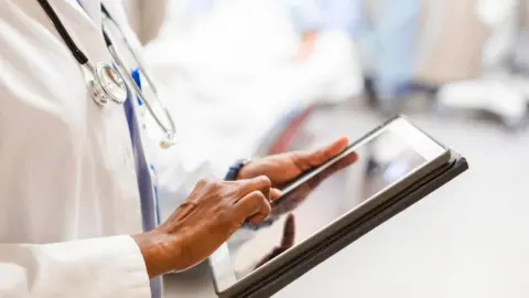 A stock image of an unrecognisable physician typing notes on the digital tablet. They are wearing a white lab coat, with a stethoscope around their neck. Items behind them are blurred. 