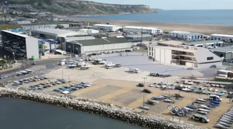 British Sailing Team Aerial view of a state-of-the-art sailing training base - a white building with balconies to the front surrounded by a boat yard with cliffs, a a beach and the sea stretching behind