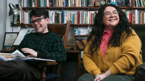 Andrew Cawley Inside the Alasdair Gray's archive, comedians Ashley Storrie and Christopher Macarthur-Boyd sit in front of a wall of bookshelves - Christopher, in a green and black patterned jumper, flips through an art book while Ashley, in green trousers, an orange shirt and a yellow cardigan, smiles off to the left.