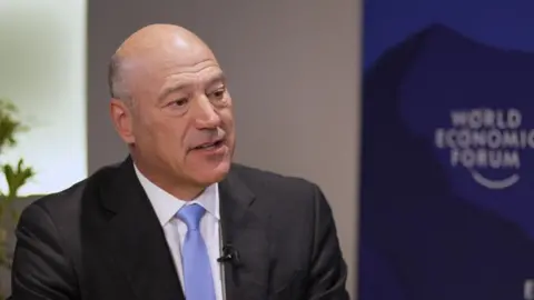 Gary Cohn wearing a dark grey suit, white shirt and pale blue tie being interviewed with a World Economic Forum sign in the background