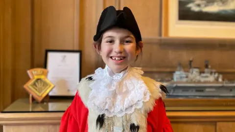 A 10 year old girl wears a red cape with a lace collar. A fur lines her shoulders. She wears a ceremonial black hat and smiles. Behind her a wooden office is blurred.