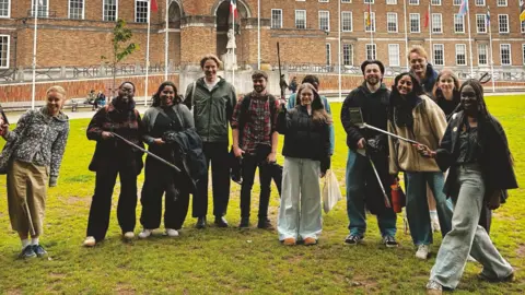 Bristol Noise/Good Faith Partnership A group of young people on a lawn in front of a large brick municipal building with a fountain in front. Bristol Several of the men and women are holding litter pickers aloft. They are all smiling to camera