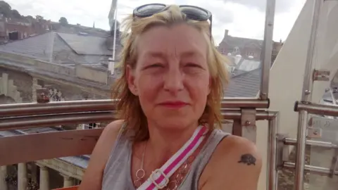 Facebook Dawn Sturgess looking at the camera. She has blonde hair and is wearing a grey vest top. She has sunglasses on her head and the strap of a bag over her shoulder is visible and is wearing a necklace. 