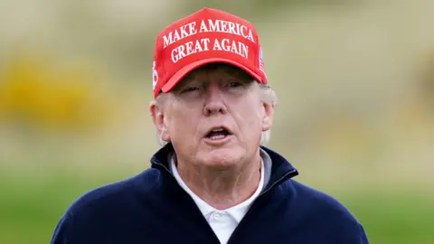 Donald Trump looking slightly off camera. He is wearing a red hat with the words 'Make America Great Again' in white text on the front. His mouth is slightly open. He is wearing a white polo shirt under a dark blue jumper which is open at the collar.