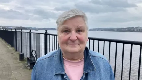 A woman with grey hair, wearing a denim shirt over a pink t-shirt. She is standing in front of railings running alongside a river.