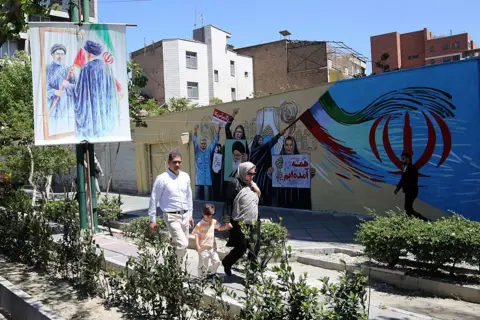 Getty Images Two adults and a small child walk along a street in Tehran which is adorned with pro-government murals 