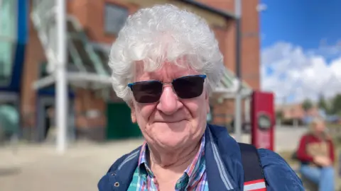 A man with curly, white hair and sunglasses. He wears a blue checked shirt and blue jacket and is stood outside a shopping centre.