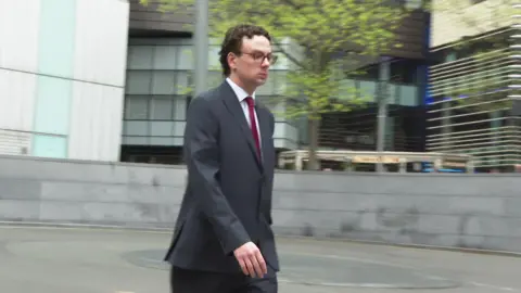 BBC Oliver Steadman arriving at Southwark Crown Court wearing a dark grey suit and a red tie. 