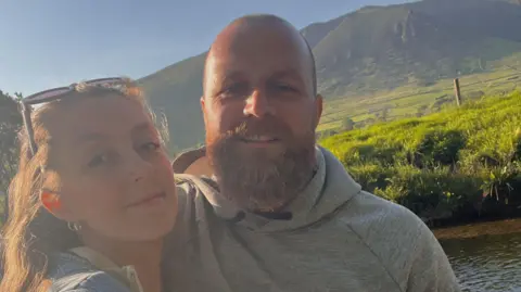 Georgia Bristow Georgia and Liam Bristow posing for a selfie together. A large green hill isvisible behind them.