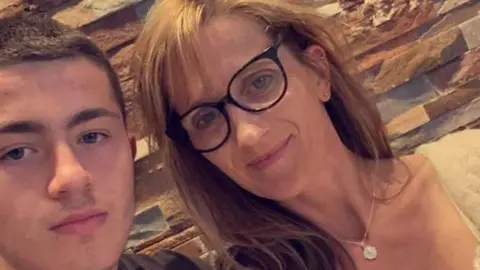 In this close up selfie image we see teenager Will Radford on the left and his mum Jane on the right. Will has short dark brown hair. Jane has long light brown hair with a fringe and wears glasses with black frames. She wears a necklace with a white pendant. 