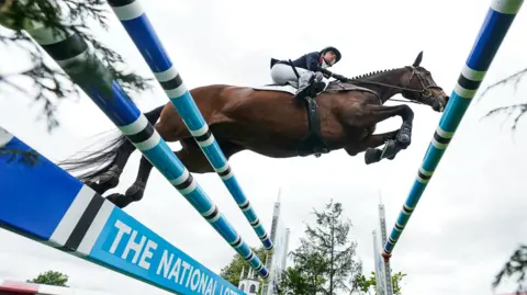 David Davies/PA Wire Rosalind Canter mid jump on her horse over blue jumps. One of them has the National Lottery written on it. Canter is wearing white jodhpurs, a dark horse riding jacket, boots and a riding hat. 