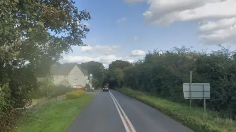 A stretch of A-road in the countryside which is bordered by a hedgerow to the right and a tree and Cotswold stone wall to the left. Further down on the left is a cream rendered pub, which has cars parked outside. Cars can be seen travelling down the road a bit further down. The sky is blue with some white clouds.