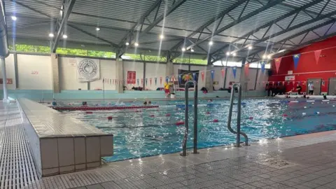 A large swimming pool with several lanes divided by red, white and blue dividers. There is a tiled wall on the left hand side, and metal handlebars on the side of the pool. People are swimming in it