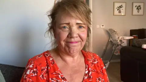 A woman in a red floral top smiling at the camera. She has blue eyes, brown hair down to her shoulders and glasses that are sat on the top of her head. In the background you see a baby high chair and two photos of plants on the wall