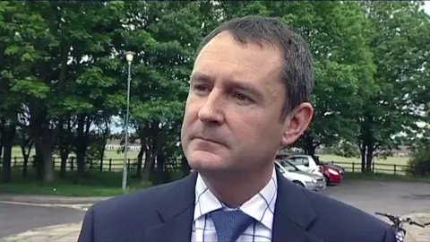 Matthew Wright is wearing a suit and looks serious as he is interviewed in a previous broadcast