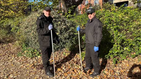 Two police officers with rubber clothes stand near a bush with sticks