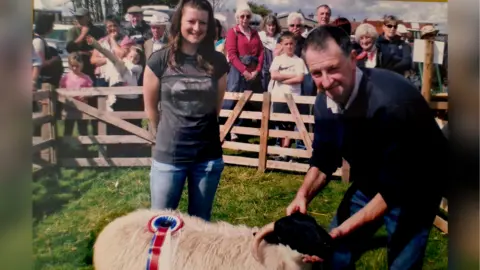 Emma Boocock An older family photo of a teenage Emma and her father standing in a fenced pen with a Scottish Blackface sheep, with a rosette on its back. The sheep has small horns, an entirely black face and white body. 